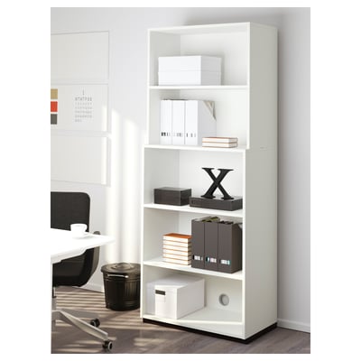 Unité de rangement de bureau blanche avec plusieurs étagères, contenant des classeurs et des boîtes, positionnée à côté d’un bureau.