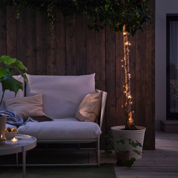 Patio confortable avec des guirlandes lumineuses GETKÅL autour de l’arbre, illuminant le mur sombre. Le canapé d’extérieur avec oreillers, corbeille de fruits et bougie ajoute de la chaleur.