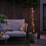 Patio confortable avec des guirlandes lumineuses GETKÅL autour de l’arbre, illuminant le mur sombre. Le canapé d’extérieur avec oreillers, corbeille de fruits et bougie ajoute de la chaleur.