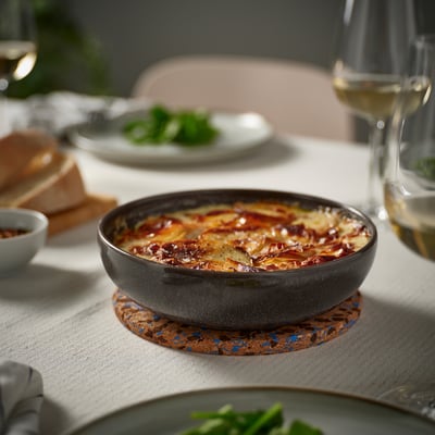 Un petit bol en grès gris foncé se trouve sur un sous-verre en liège, rempli de plats superposés. Entouré d’assiettes et d’un verre à vin, sur une nappe légère.