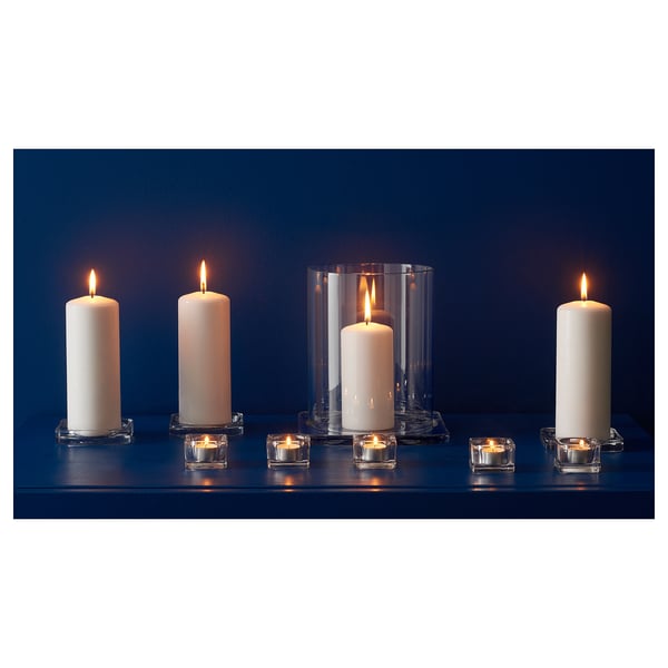Quatre grandes bougies blanches dans des supports en verre transparent de différents motifs sur une étagère bleu foncé.