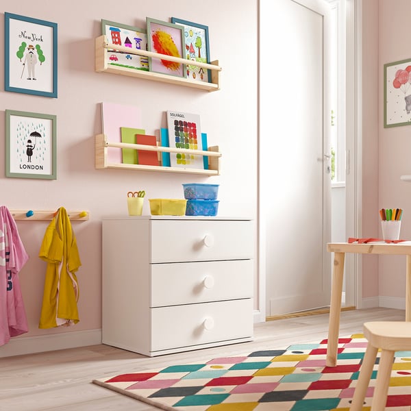Chambre d’enfant avec commode blanche à 3 tiroirs, étagères remplies de jouets, murs roses.