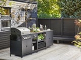 Configuration moderne de la cuisine extérieure : gril en métal noir, comptoir avec des légumes et étagères sur terrasse en bois.