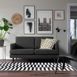 Salon moderne avec canapé GRILLSTORP noir, oreillers gris, tapis à carreaux noir et blanc, art mural et plante.