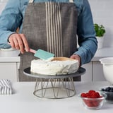 Personne glaçage gâteau avec une spatule bleue sur un support métallique avec des framboises et des myrtilles dans des bols à proximité.