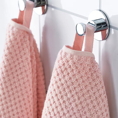 Deux serviettes rose clair pendent soigneusement à des crochets chromés sur une porte blanche. La surface douce et texturée des serviettes indique l’absorption et le confort.