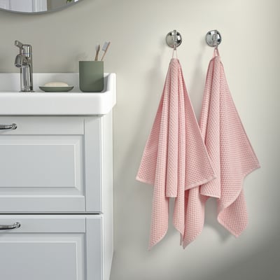 Deux serviettes GULVIAL roses sur crochets, douces et texturées, à côté d’un meuble-lavabo blanc propre avec une tasse verte, des brosses à dents et du savon.