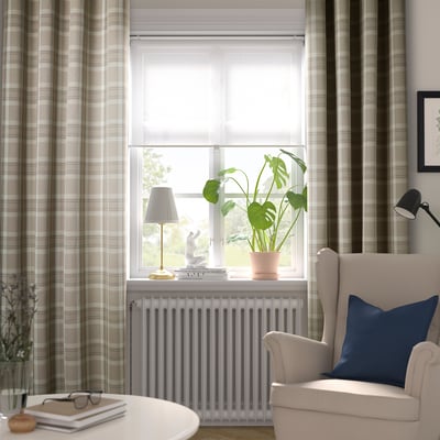 Salon lumineux avec de grands rideaux HÄGGVECKMAL beiges, une chaise douillette et une plante en pot près d’une fenêtre, offrant une atmosphère détendue.
