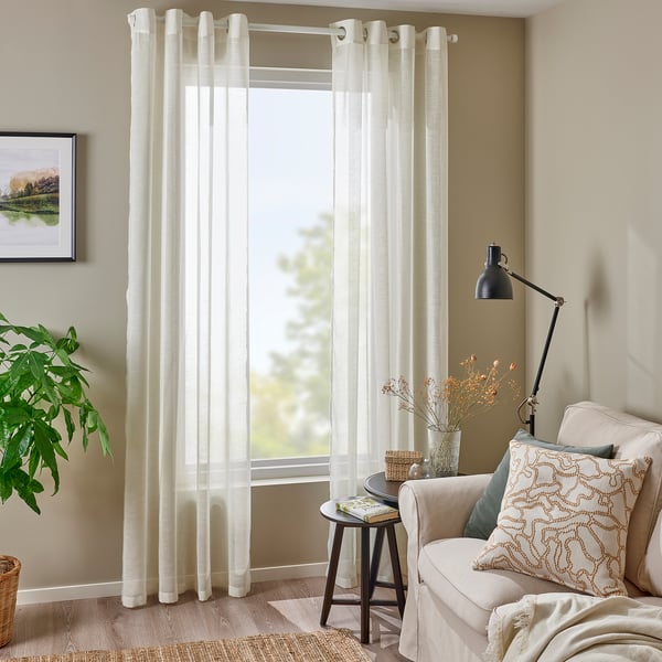 Salon lumineux avec rideaux transparents blancs HÄLLEBRÄCKA, chaise beige, lampadaire noir et plante verte.