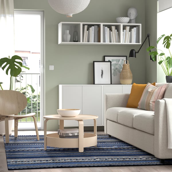 Salon moderne avec murs verts, tapis bleu, canapé blanc, table basse en bois, buffet, plantes, lampe et porte de balcon.