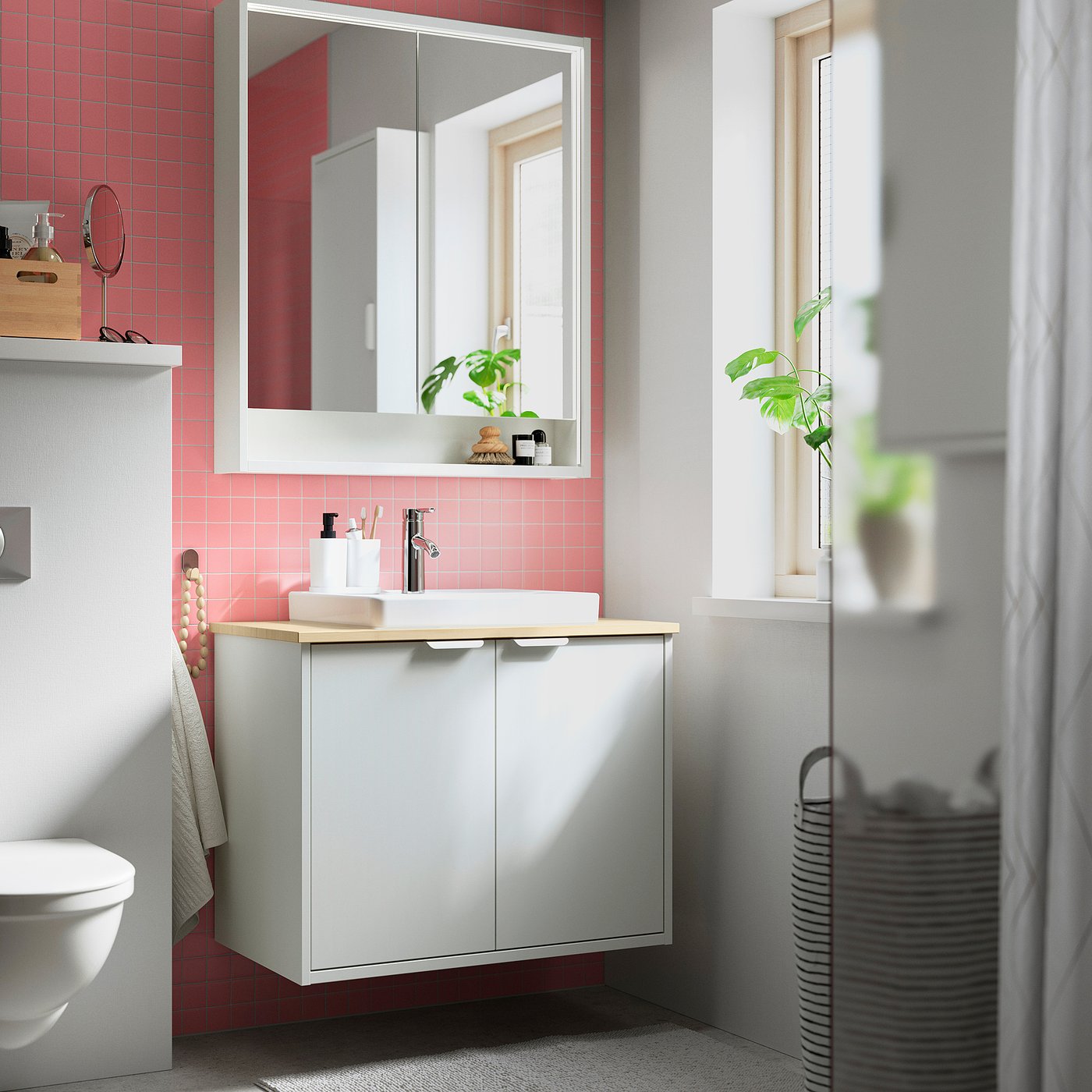 Salle de bain moderne : meuble-lavabo HAVBÄCK blanc, carrelage rose, grand miroir, comptoir en bois, plante verte, panier gris, espace lumineux.