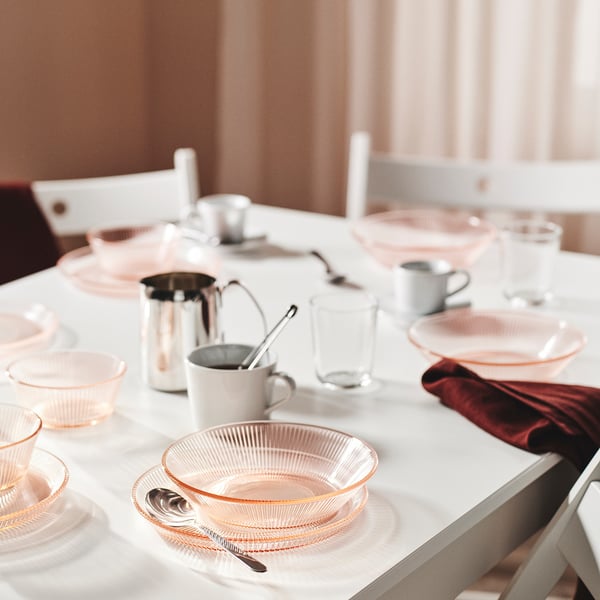 Ensemble de vaisselle en verre rose sur une table blanche, avec des assiettes, des bols, des tasses et des couverts, présentant un design élégant et une durabilité.