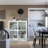 Meuble de rangement HAVSTA blanc avec portes vitrées, bois de pin et verre trempé, exposant vaisselle et livres, sous une horloge noire.