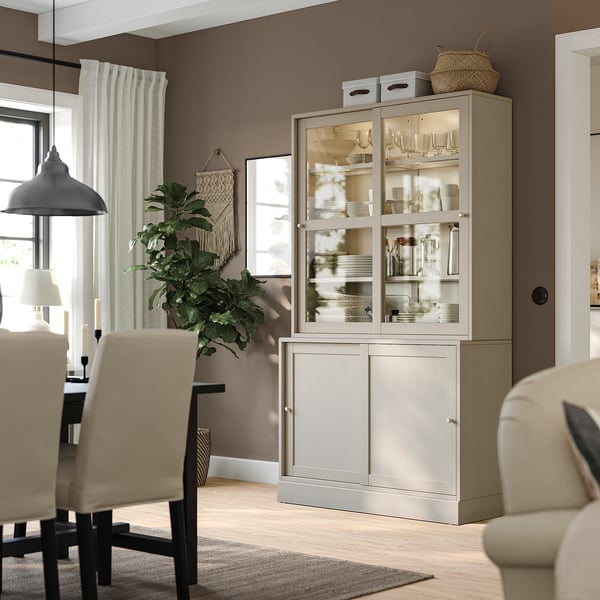 Une armoire HAVSTA gris clair avec des portes vitrées, exposant de la vaisselle et des verres, dans une salle à manger avec des chaises beiges, une déco végétale et bohème.
