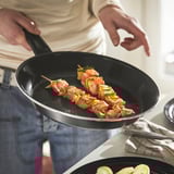 La personne fait cuire des brochettes de viande et de légumes dans une poêle antiadhésive avec une poignée noire, adaptée à tous les types de plaques de cuisson.