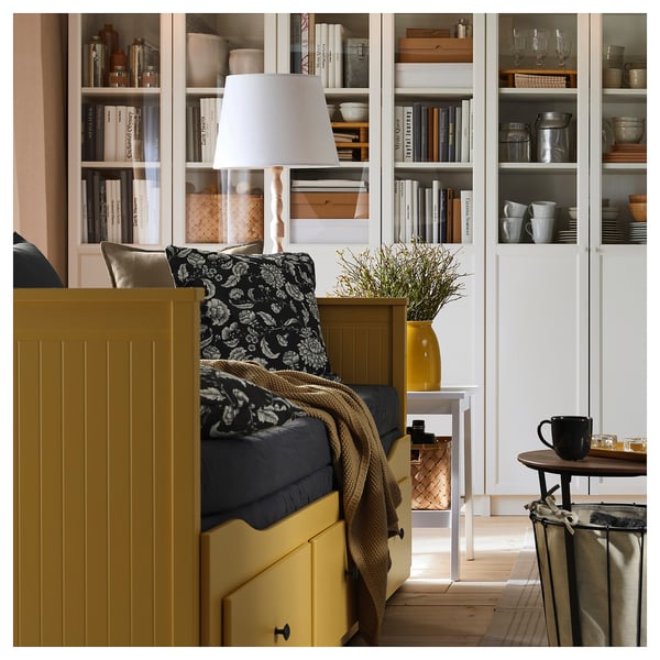 Une méridienne jaune avec des tiroirs, des oreillers floraux, une lampe et des étagères avec des livres et des tasses.