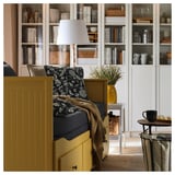 Une méridienne jaune avec des tiroirs, des oreillers floraux, une lampe et des étagères avec des livres et des tasses.
