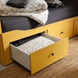 Lit de rangement jaune vif avec tiroir ouvert, montrant une literie florale à l’intérieur. Idéal pour les espaces compacts, combinant fonctionnalité de rangement et de sommeil.