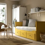Lit de jour HEMNES jaune vif avec tiroirs de rangement, oreillers moelleux et étagères intégrées à côté d’une armoire blanche, près d’une fenêtre avec une chaise en osier.