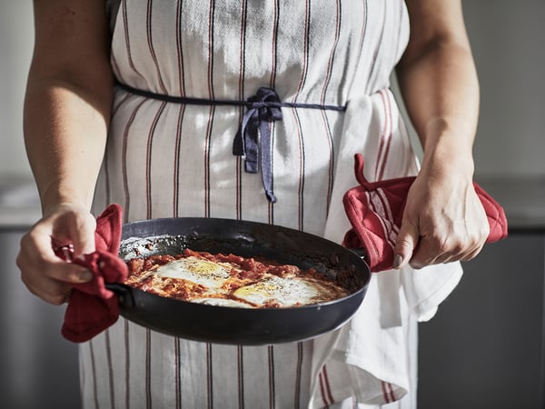 Personne tenant une poêle avec des aliments frits, portant un tablier et des mitaines rouges.