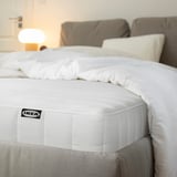 Lit soigné avec matelas ikea blanc, literie grise, canapé, lampe en arrière-plan.