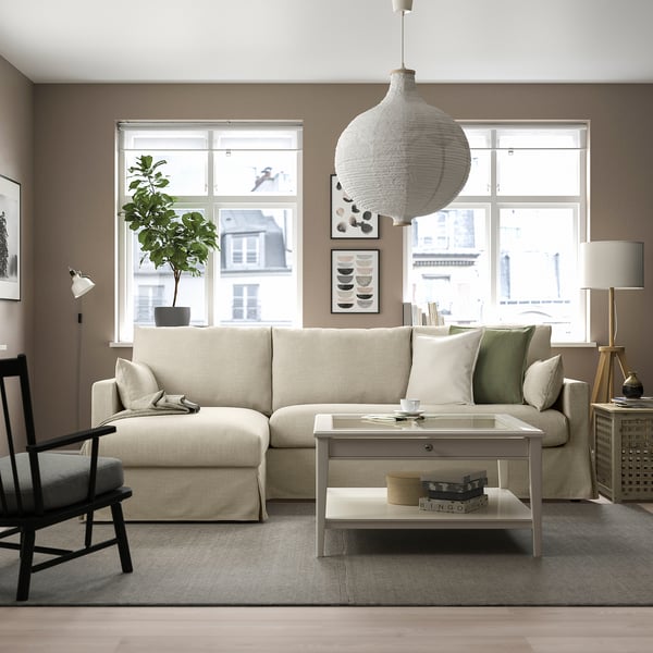 Salon moderne avec canapé crème HYLTARP, rangements et table basse blanche. Grandes fenêtres, tapis beige, chaise noire/blanche et suspension.