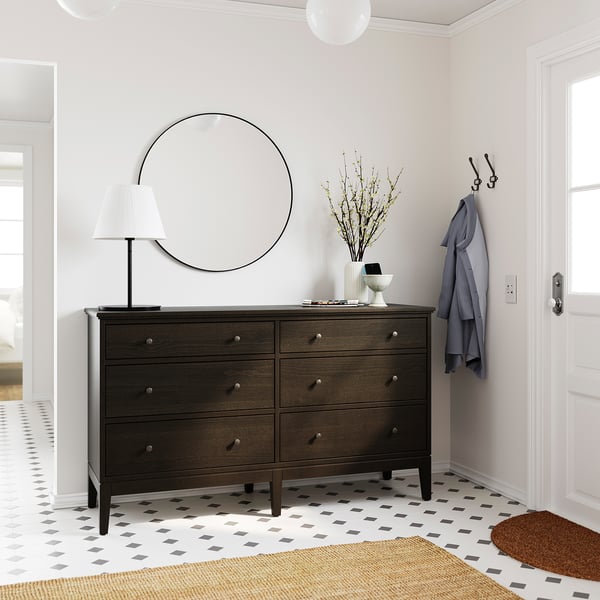 Chambre moderne avec commode à 6 tiroirs marron foncé IDANÄS, miroir rond, lampe, branches décoratives et patères.