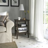 Table d’appoint IDANÄS à côté d’une chaise grise, tenant une lampe, des tasses, des livres. Bois sombre, sols blancs. Idéal pour les espaces restreints.