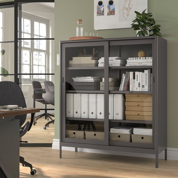 Armoire de rangement noire IDÅSEN avec portes vitrées dans le bureau, contenant des livres et des objets de décoration, économise de l’espace.