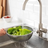 Personne lavant des légumes verts dans une passoire en métal sous un robinet de cuisine en argent.