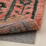 Tapis tissé coloré sur un plancher en bois avec un sous-tapis gris visible en dessous.