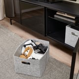 Boîte de rangement en feutre près de l’unité TV, contenant diverses manettes de jeu, avec des poignées découpées pour un transport facile et une organisation soignée.