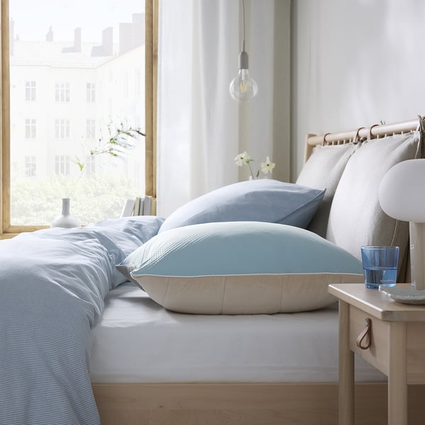 Lit avec protège-oreiller bleu, draps blancs, cadre en bois, verre d’eau, lampe, vue sur la ville.