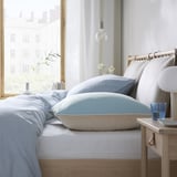 Lit avec protège-oreiller bleu, draps blancs, cadre en bois, verre d’eau, lampe, vue sur la ville.