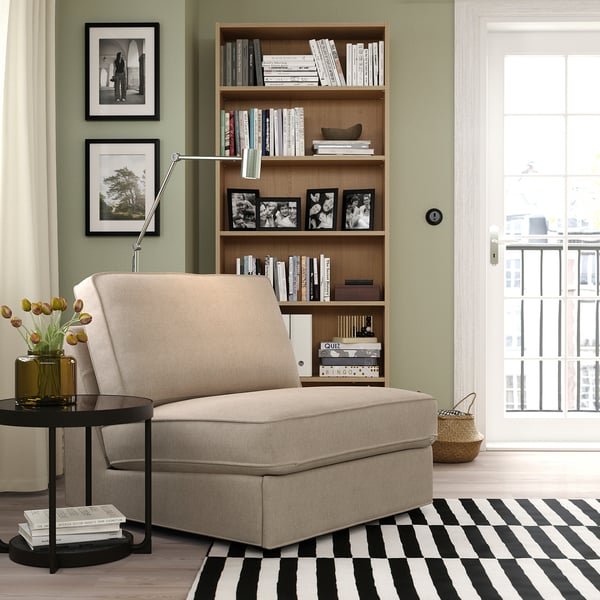 Salon moderne avec des murs en sauge, un canapé-lit KIVIK beige, un tapis rayé et une étagère en bois remplie de livres et de photos en noir et blanc.