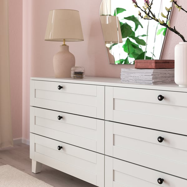 Commode blanche avec six tiroirs, boutons noirs, lampe et livres.