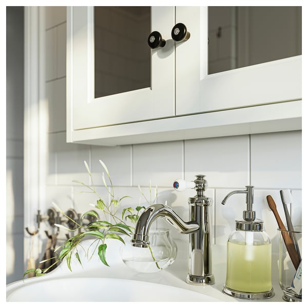 Salle de bain moderne avec robinet chromé, distributeur de savon et plante en bulle de verre. Dosseret et armoire en tuiles blanches avec boutons noirs.