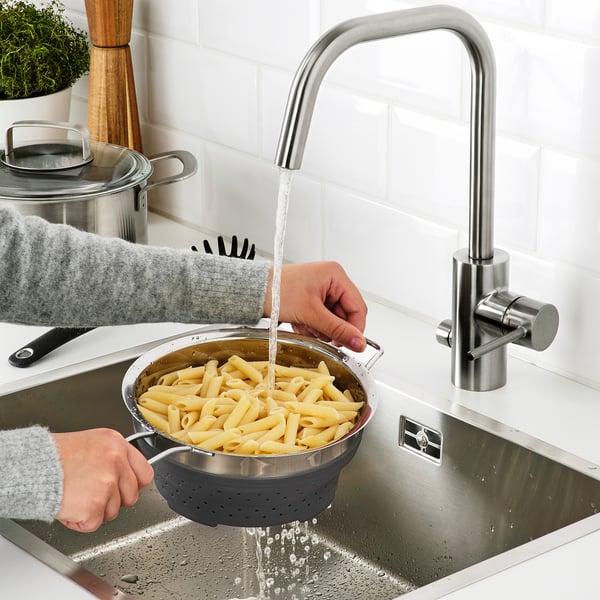 Personne rinçant des pâtes cuites dans un évier en acier inoxydable à l’aide d’une passoire en silicone flexible avec de l’eau courante.