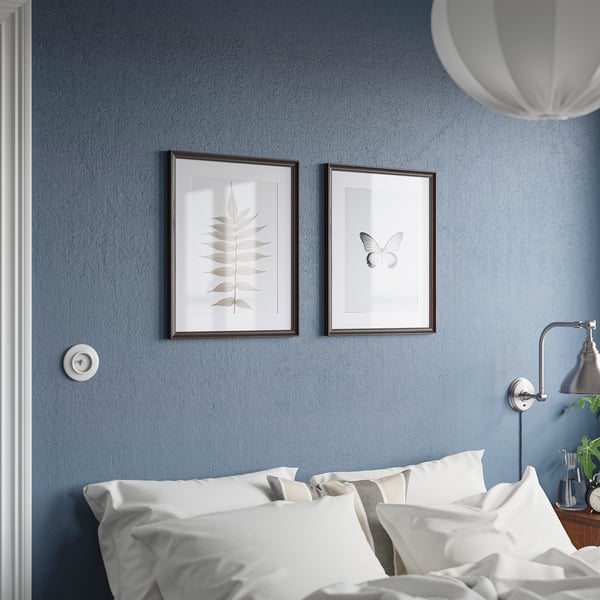 Chambre bleue luxueuse avec literie blanche et deux cadres KLOTKÖRSBÄR affichant des imprimés de feuilles et de papillons sur les murs bleu marine, soignée et élégante.