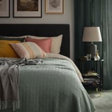Chambre à coucher avec couvre-lit KÖLAX gris-vert, coussins à motifs, table de chevet avec lampe, livres et œuvres d’art encadrées au mur.