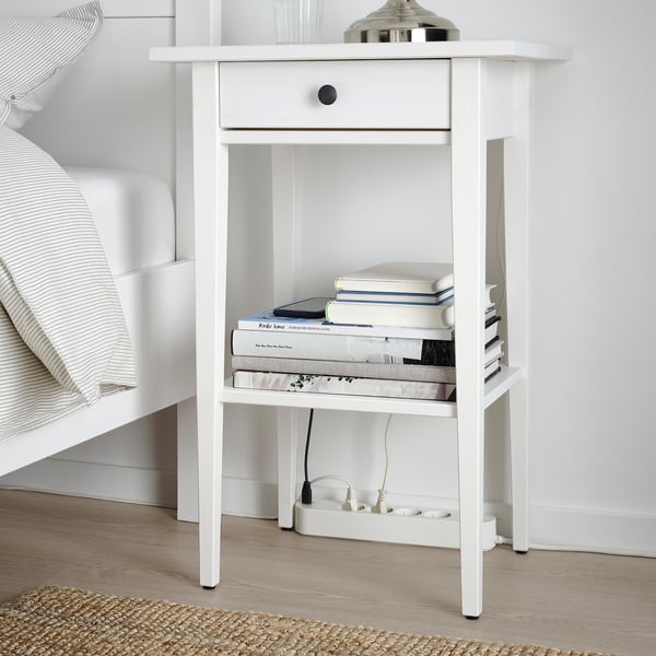 Table de chevet blanche avec un seul tiroir et une étagère ouverte, contenant des livres et une lampe avec des capacités de chargement USB.