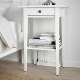 Table de chevet blanche avec un seul tiroir et une étagère ouverte, contenant des livres et une lampe avec des capacités de chargement USB.