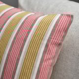Gros plan d’une housse de coussin rayée avec un tissu doux et texturé. Rayures beige, rouge clair et gris, avec un éclat subtil. Fermeture éclair près du bord.