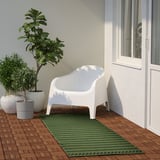 Tapis vert KORSNING dans le coin salon. Rayures lumineuses sur parquet, chaise blanche, deux grandes plantes à proximité. Dimensions des tapis : 96 x 26.