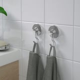 Deux crochets à ventouse argentés accrochent des serviettes grises sur un mur carrelé blanc, offrant un rangement organisé dans la salle de bain sans avoir besoin de percer.