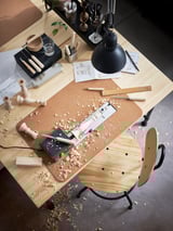 Un espace de travail animé : bureau en bois clair KULLABERG parsemé d’outils de bricolage, de copeaux de bois, de croquis et d’une lampe noire.