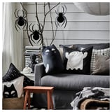 Salon sur le thème d’Halloween avec canapé gris, décoré avec des coussins noirs et blancs KUSTFYR. Des araignées noires pendent au-dessus, créant une atmosphère festive.