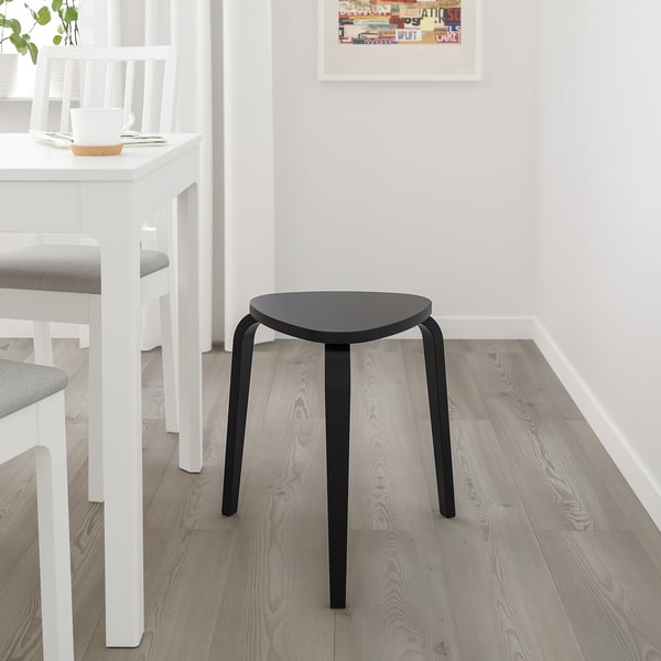 Tabouret KYRRE : noir élégant, assise triangulaire, trois pieds courbés. Petit, se glisse sous une table blanche, près d’une chaise rembourrée grise.
