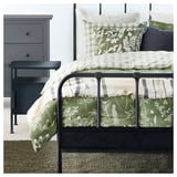 Une chambre douillette avec une parure de lit verte et blanche et une commode grise. Un tapis en jute vert foncé est sur le sol.