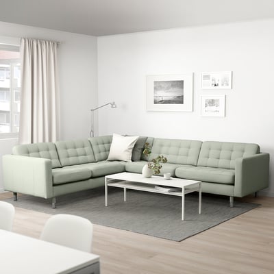 Salon minimaliste moderne : canapé en forme de L vert clair, table basse blanche, tapis gris, art noir et blanc, grande fenêtre, lampadaire.